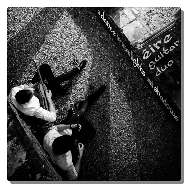 Black and white cover art for A Dance Between Lights And Shadows by Éire Guitar Duo featuring two classical guitarists seated on gravel in dramatic overhead lighting.
